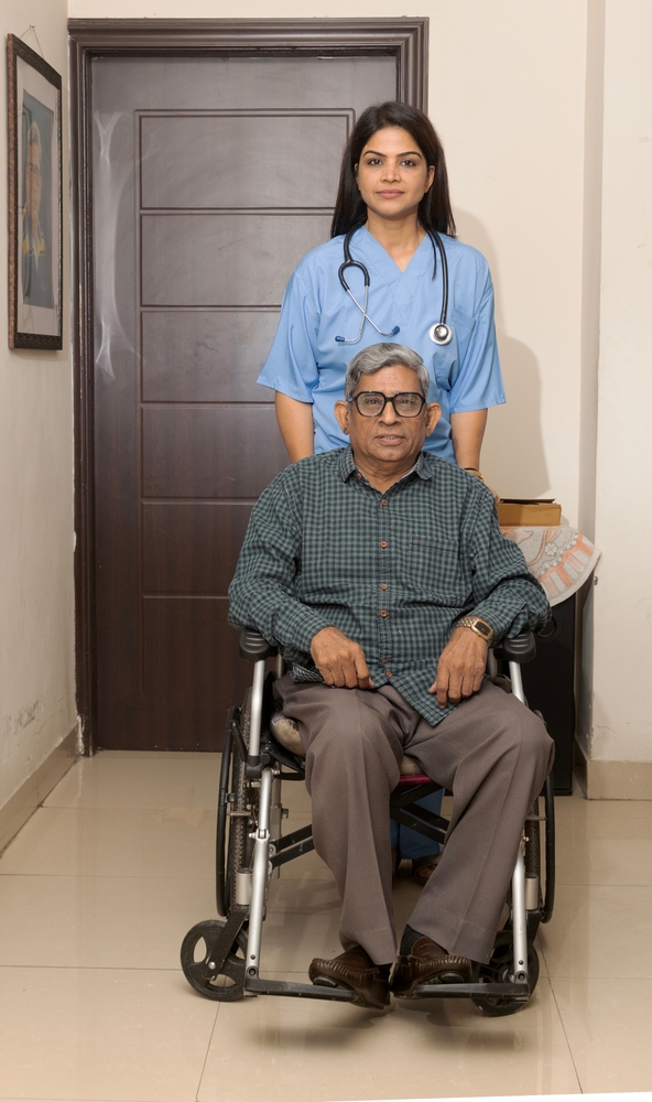 A caregiver standing behind an elderly man seated in a wheelchair.